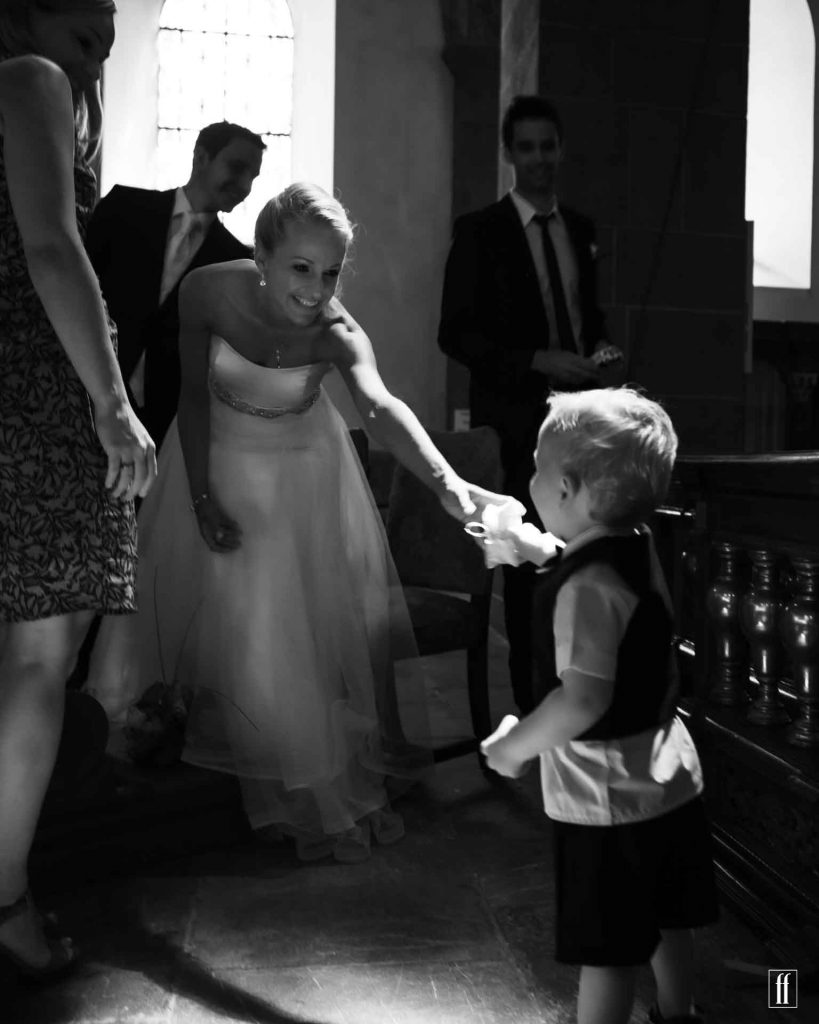 Hochzeitsfotografie einer Braut, die von einem kleinen Kind in Weste und Shorts ihren Ehering gereicht bekommt. Die Braut lacht, der Bräutigam und der Trautzeuge schauen ebenfalls lachend auf das Geschehen.