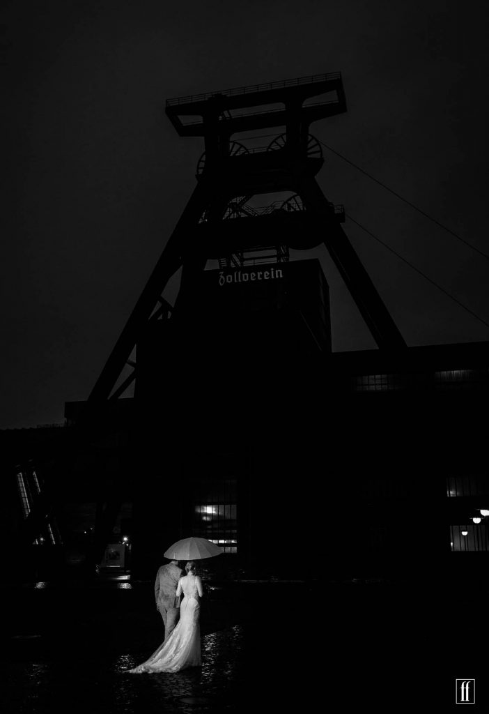 Braut und Bräutigam spazieren im dunkeln bei Regen vor dem großen Turm der Zeche Zollverein