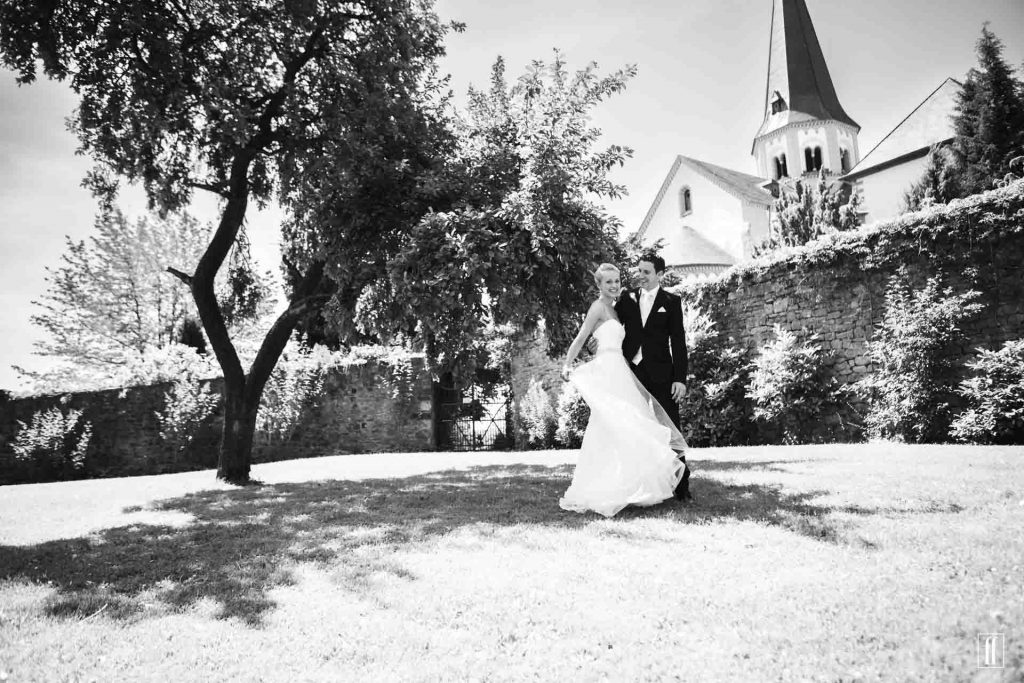 Hochzeitsfotografie in der Eifel. Ein Brautpaar steht in einem Klostergarten unter einem Baum und lacht. Im Hintergrund ist der Turm der Basilika zu sehen.